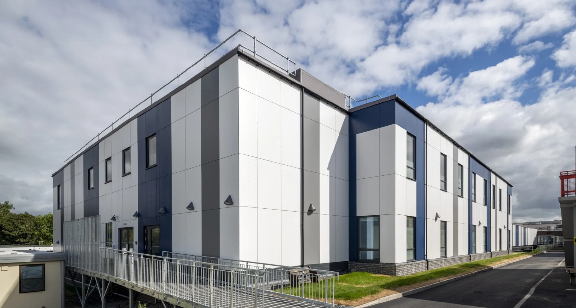 External three-quarter view of the two-storey hospital building at Airedale. The external classing is white with blue and grey stripes and there are windows at regular intervals on both floors. To the left is the building entrance, with a long, gentle ramp running up to the doors. In the foreground and leading off to the right is a tarmac walkway. The sky is blue and dotted with white clouds.
