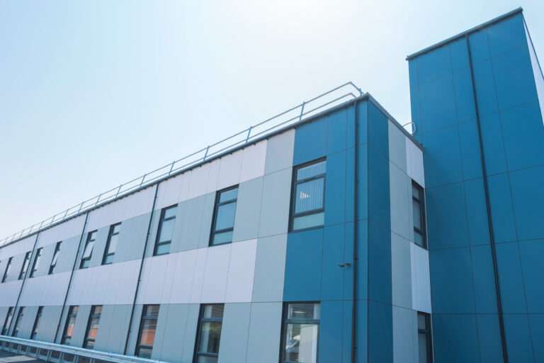Exterior of the new ward building at Leighton Hospital. The cladding is a mix of blue, grey and white. The main building is two storeys, with a three storey staircase and elevator tower to the right. There are regular windows on each floor. The sky is clear blue.
