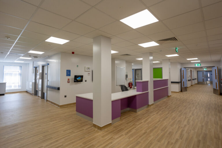 A wide corridor incorporating a nurses' station. The walls and ceiling are white, while the nurses' station is finished in purple. To the left there is a large window, to the right the corridor passes through three sets of light blue double doors. The flooring is a beech colour wood effect vinyl.