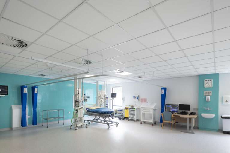A high dependency ward room, featuring a hospital bed and a lot of medical monitoring equipment. To the right, there is a desk and chair with computer equipment on the desk. Next to this is a hand washing station. In the background there is a large window, and the rear wall is a light blue colour.