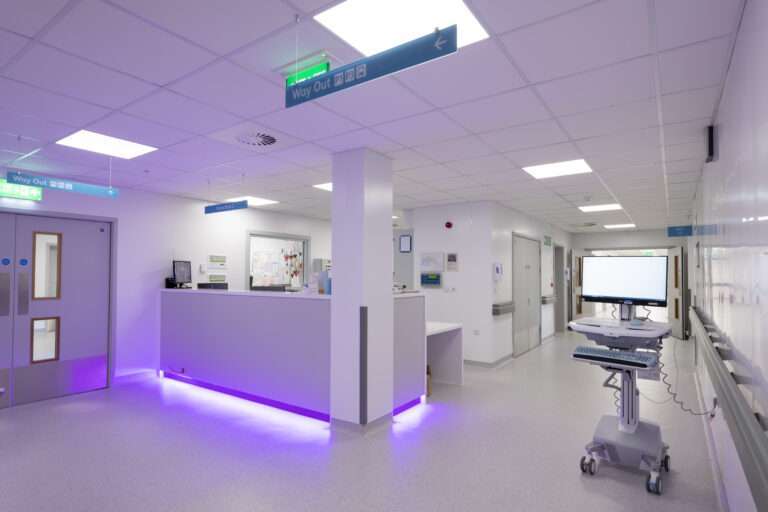 A hospital corridor featuring a nurses' station. The walls are white, there is overhead lighting and the station features purple ambient lighting. To the left is a set of double doors, to the right the corridor continues through another set of doors and there is a trolley containing some medical equipment in the foreground.