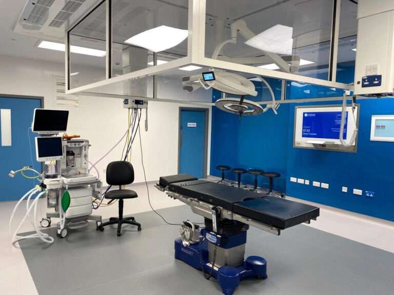 A new operating theatre. The rearmost wall is blue and features a couple of digital screens. In the foreground there is an operating table with lights over the top, as well as a number of items of medical equipment.