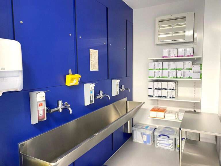 A scrub room for an operating theatre. To the left, there is a blue wall with a large stainless steel washing trough along it, with taps and soap dispensers on the wall. To the right, there is a bank of shelves stocked with medical supplies.