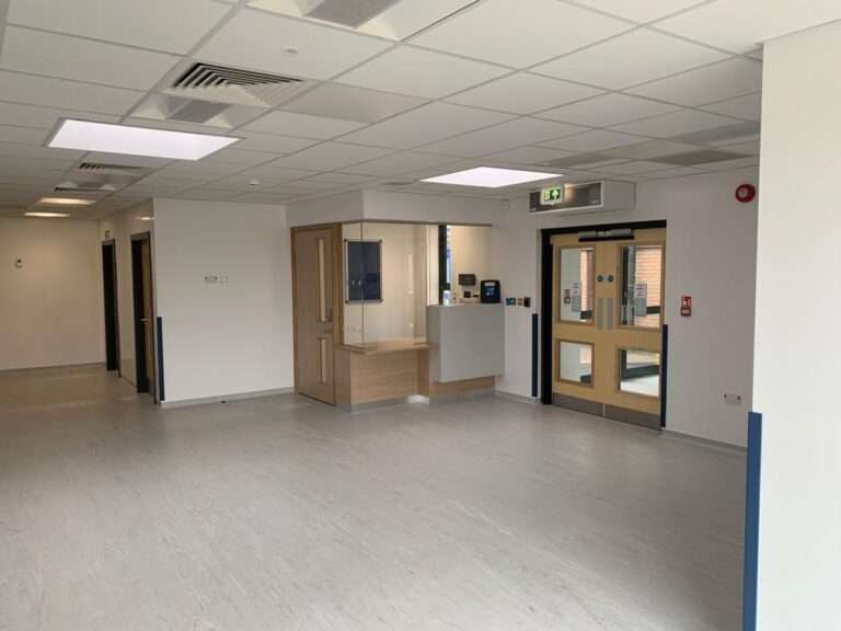 Inside the reception area of the SDEC. The walls and ceiling are white. The floor is a light grey vinyl. In the far corner there is a booth for the receptionist, sealed behind glass and a wooden door. To the right hand side is the main entrance which is a double door.