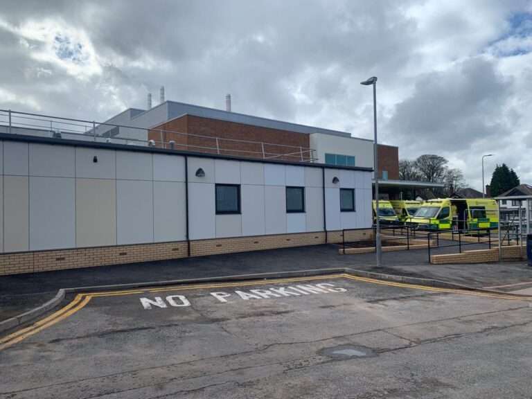 Exterior of the SDEC building. The lower section is finished in brick, the top three-quarters is clad in white and cream panels. To the right there are a number of ambulances parked. The sky is cloudy and grey.