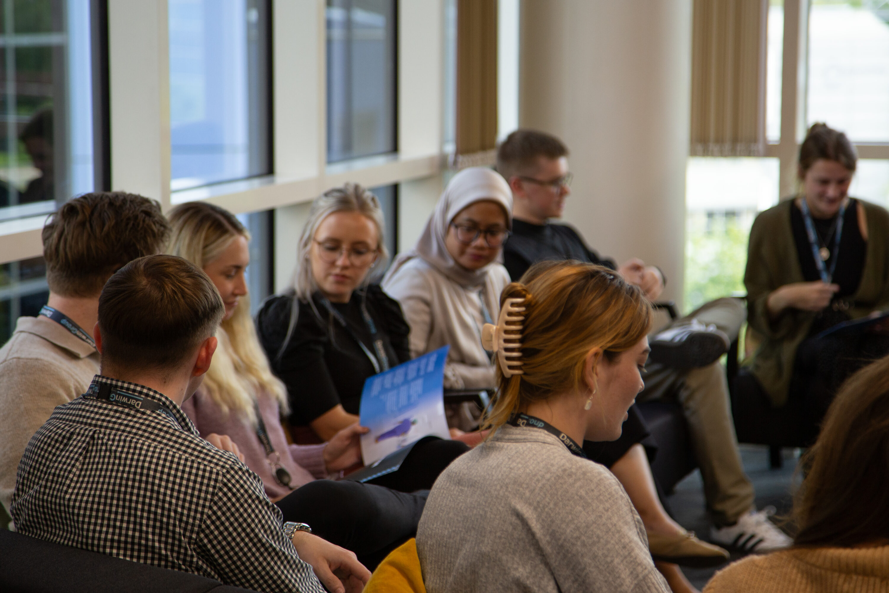 A group of Darwin Group employees sat in the staff hub, they are looking through a brochure and there are large windows in the background.