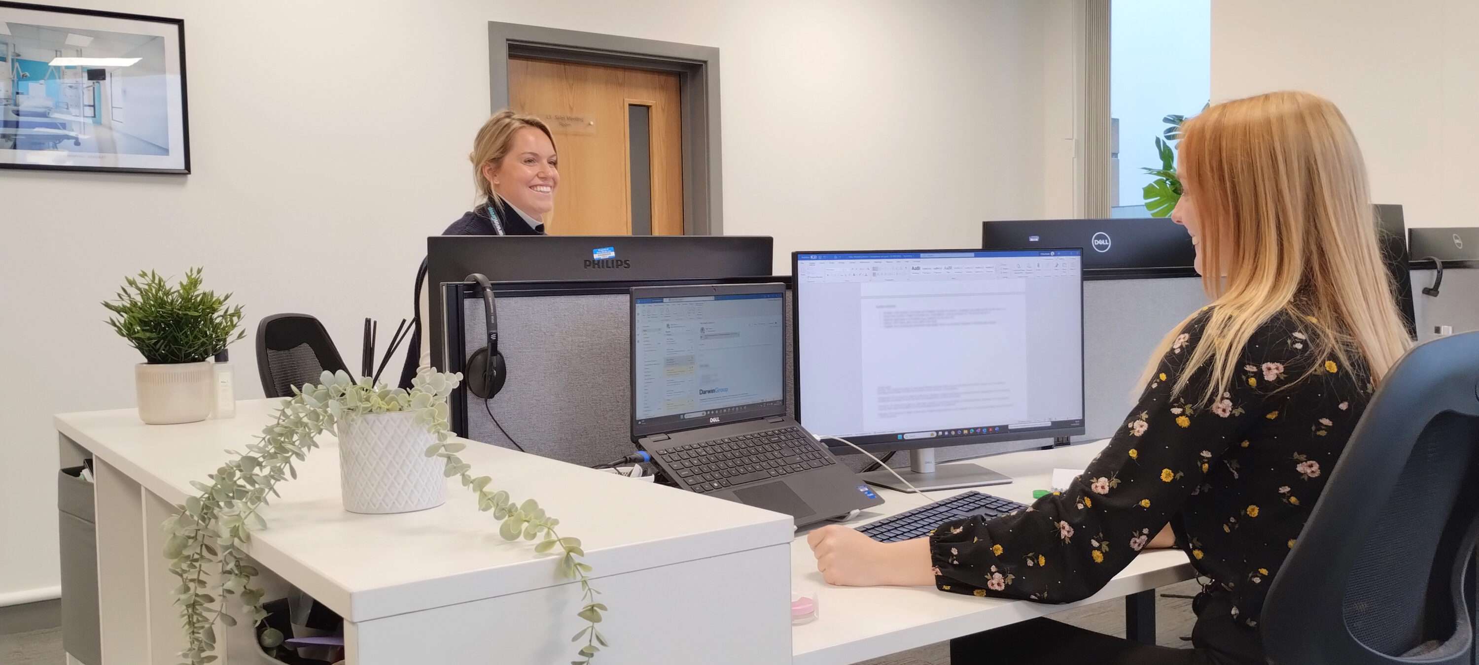 Two female Darwin Group employees, sat on opposite sides of a desk with computer equipment in between. They are both smiling.