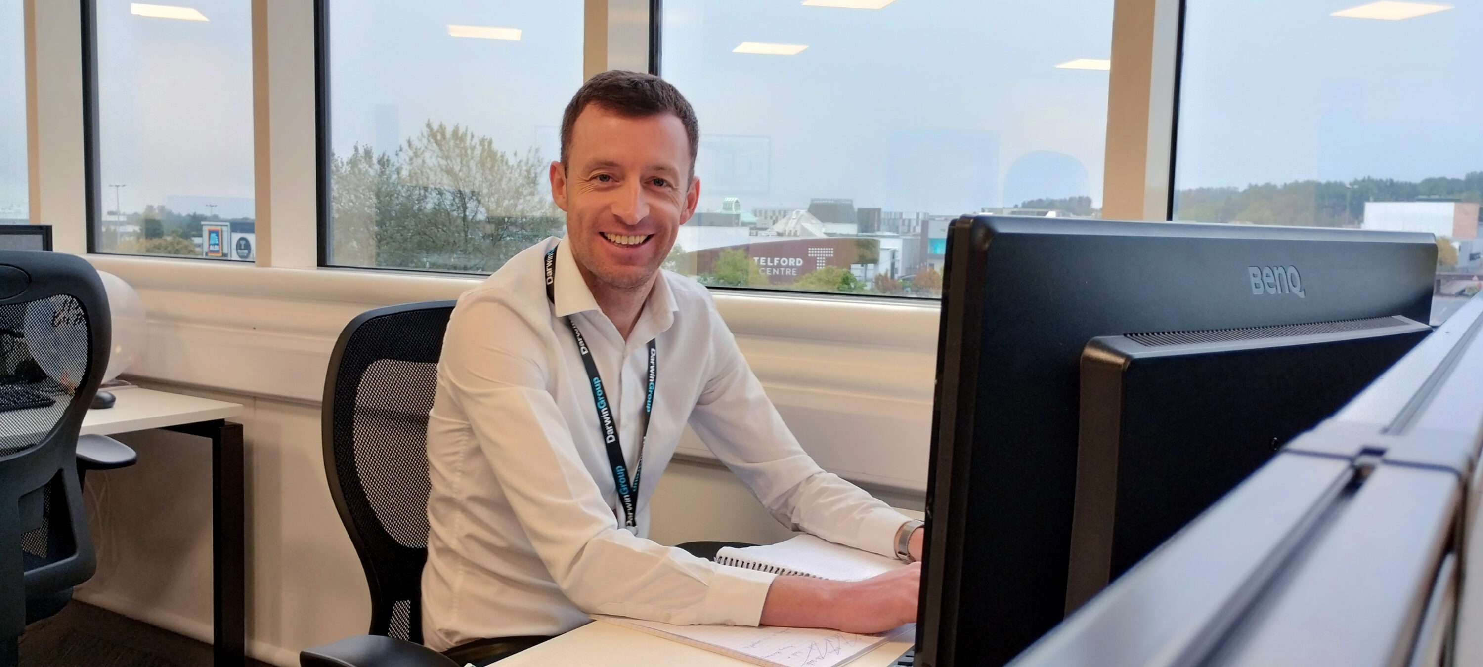 A Darwin Group employee, sat at their desk, looking at the camera and smiling. In the background you can see out of the office windows.