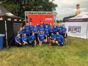 The Darwin Group team, ready to start the Tough Mudder challenge. There is a large banner behind them, gazebos to either side, the sky is overcast and they are stood in a grassy field.