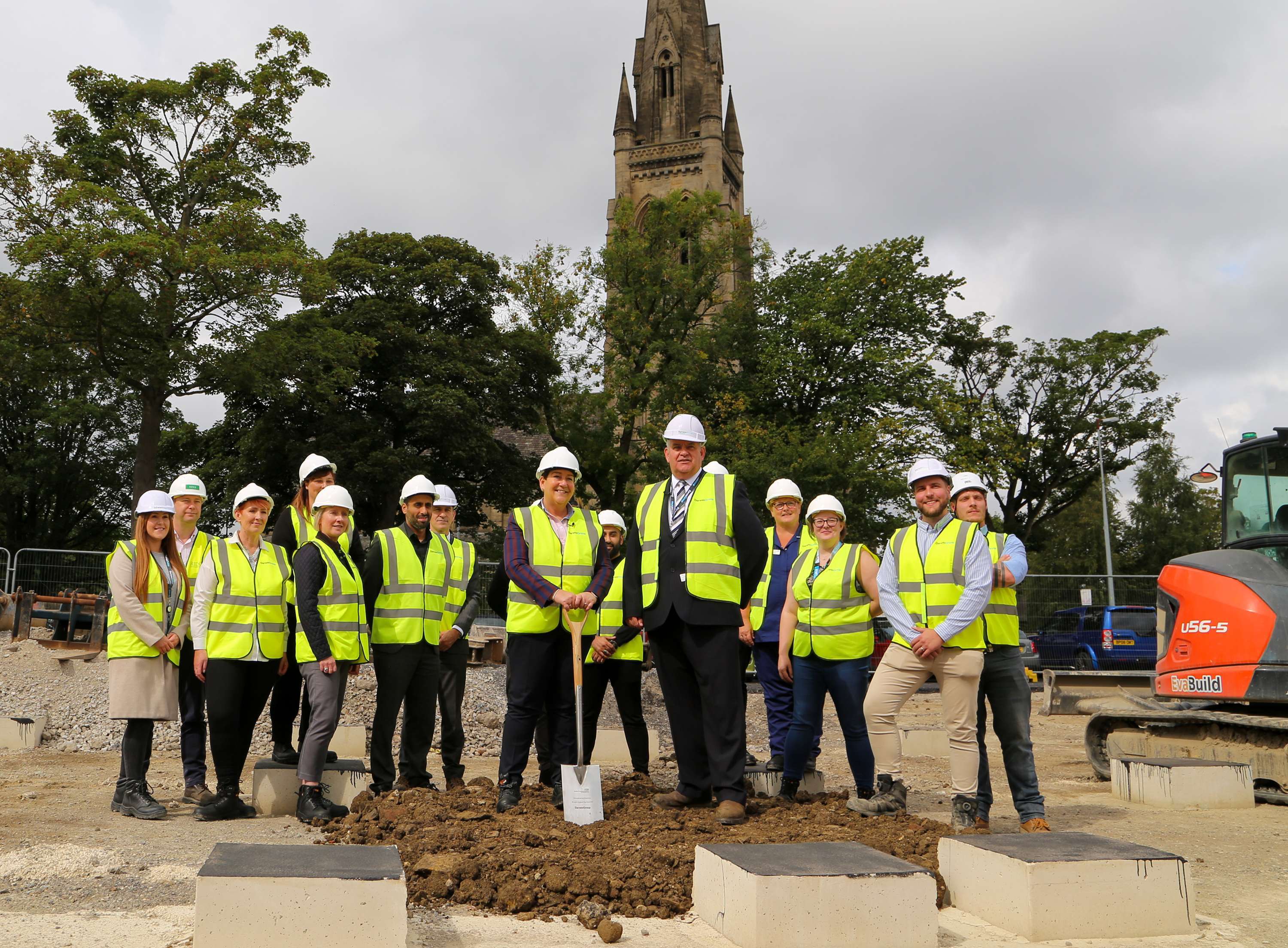 A group of Darwin Group staff and clinicians at the official ground breaking ceremony for St Luke's in Bradford. In the centre foreground there is a woman stood holding the ceremonial spade. In the background there is a line of trees and the spire of a church.
