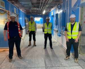 Four men in workwear with high-visibility vests and hard hats. They are standing in the corridor of an under-construction hospital building.