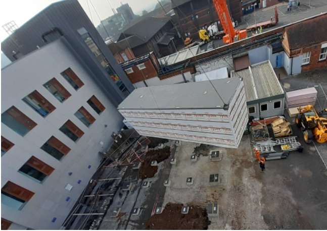 A bird's eye view of a construction site. The groundworks have been completed and a module is being craned into place alongside the existing hospital building.