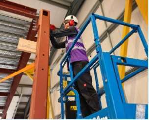 A production worker wearing full PPE and standing in a cherry-picker cradle. They are working on the construction of a steel frame.
