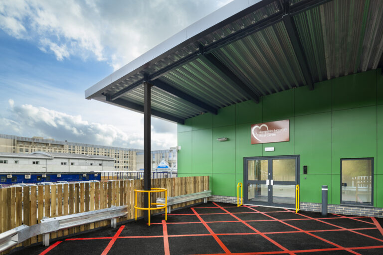 The exterior of Basingstoke Cardiac Catheterisation Labs. The cladding is greed, and the curved roof is made from steel. In the background you can see the existing older hospital buildings. The sky is blue with lots of white clouds. In the foreground, there is a large area of hashed red paint in front of the building entrance.