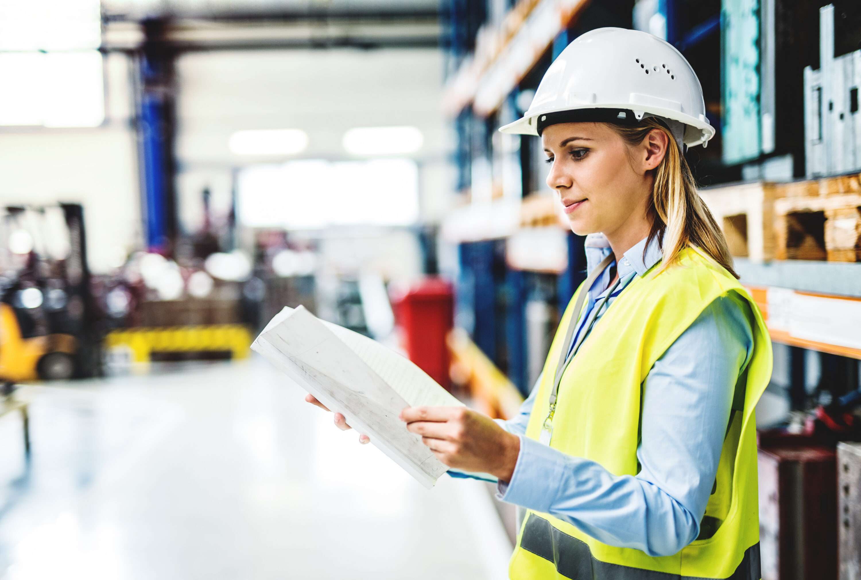 A woman in high-visibility jacket and hard hat, examining some paperwork while standing in a factory or warehouse facility.