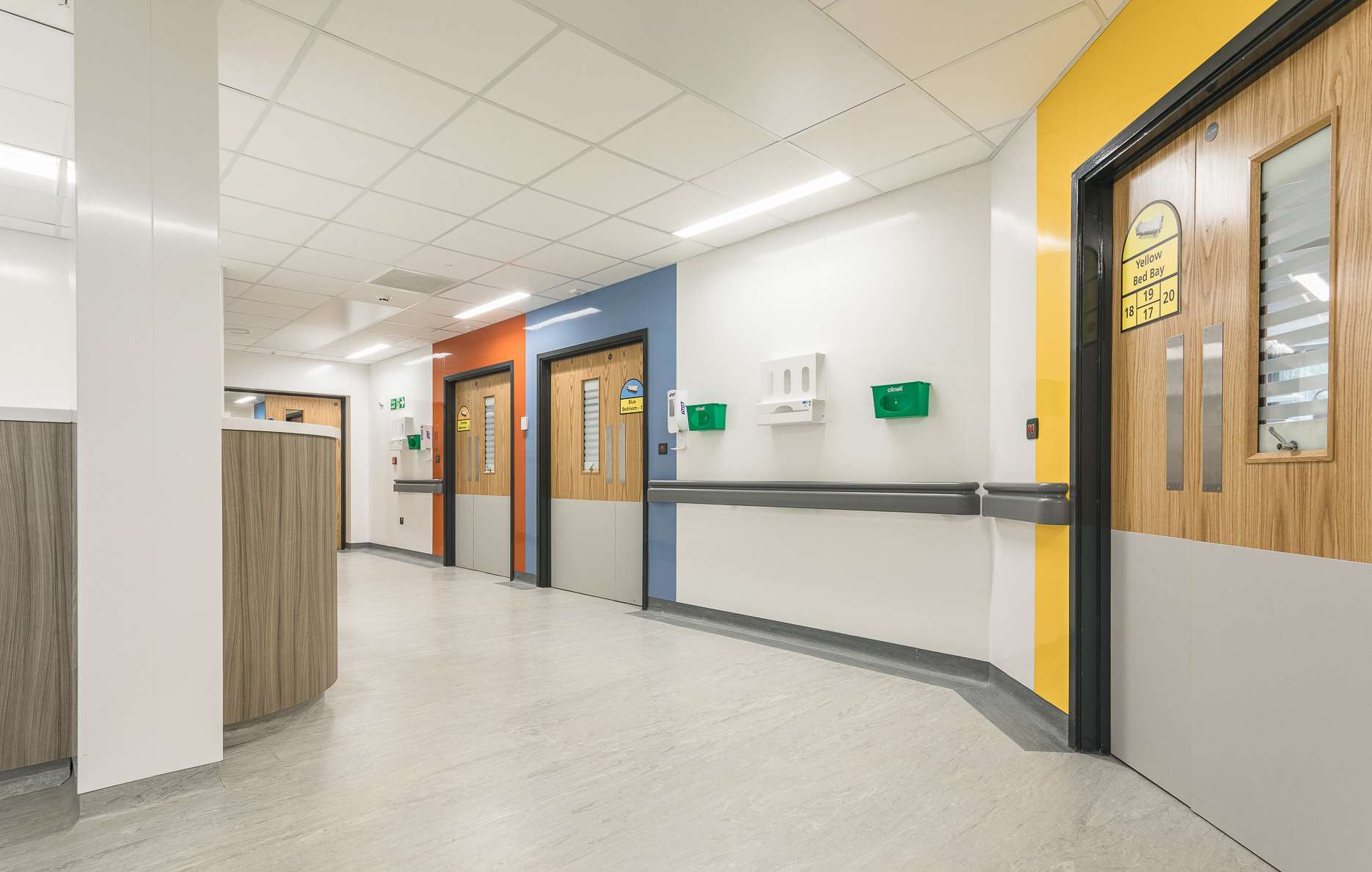A light hospital corridor, finished mostly in white and curving around to the left. To the right there are three sets of double doors, each with a different colour surround - yellow, blue and orange.