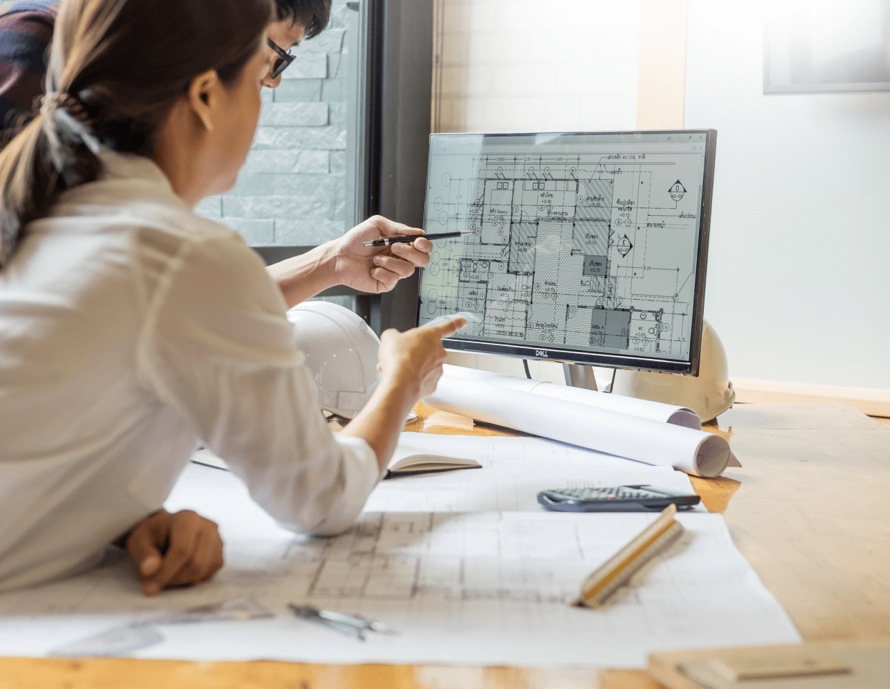 A woman and a man in an office, sitting at a desk and pointing at a computer screen, which is displaying a set of technical plans for a building. On the desk in front of them are a number of printed out plans, some open and some rolled up.