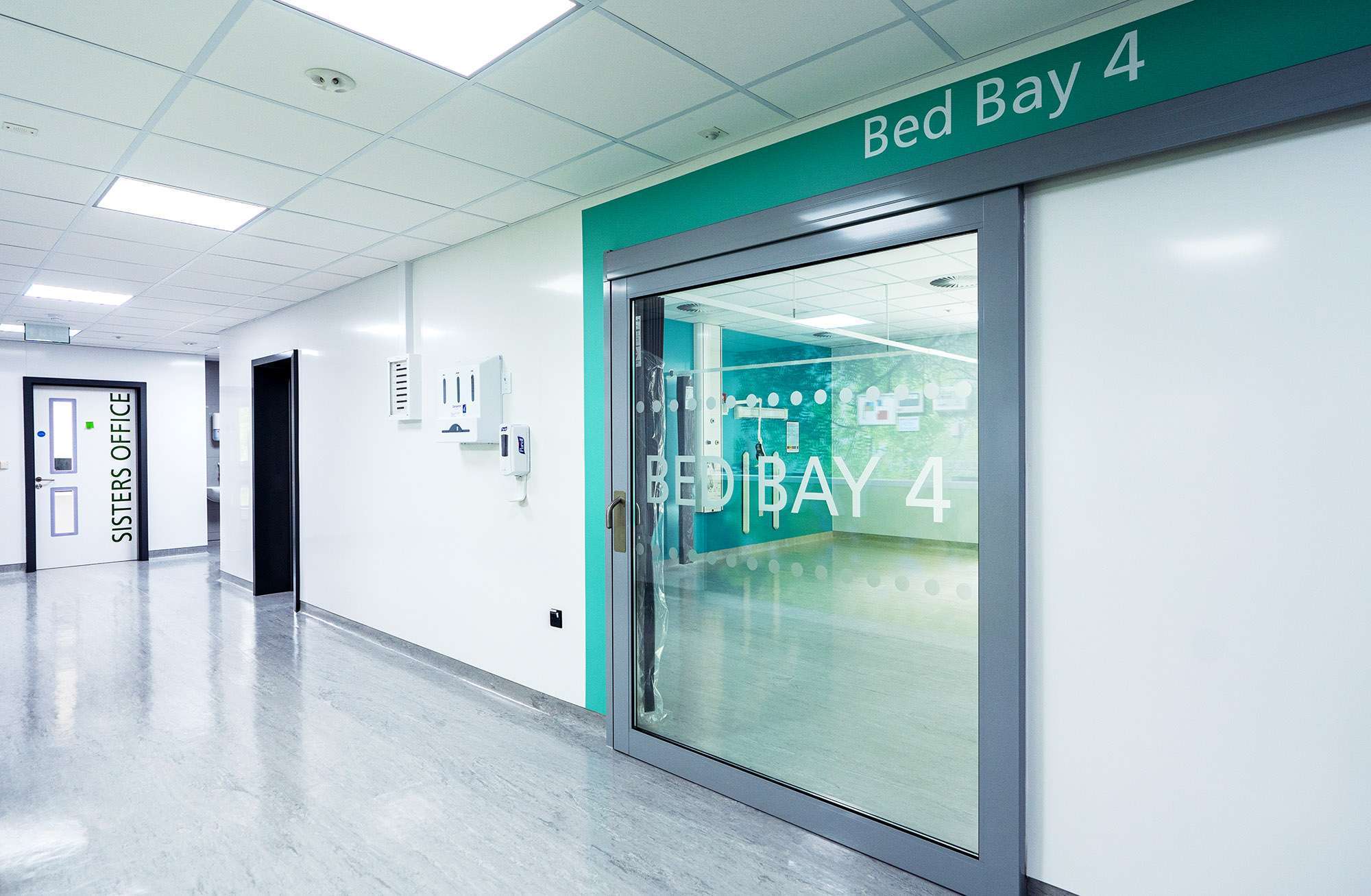 A white hospital corridor, with a large glass door leading to a bay for beds. The surrounding area of the door is green.