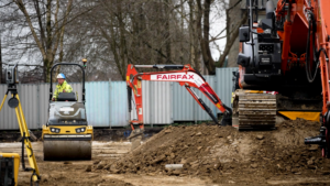 A small digger and a roller on a building site, with site cabins in the background