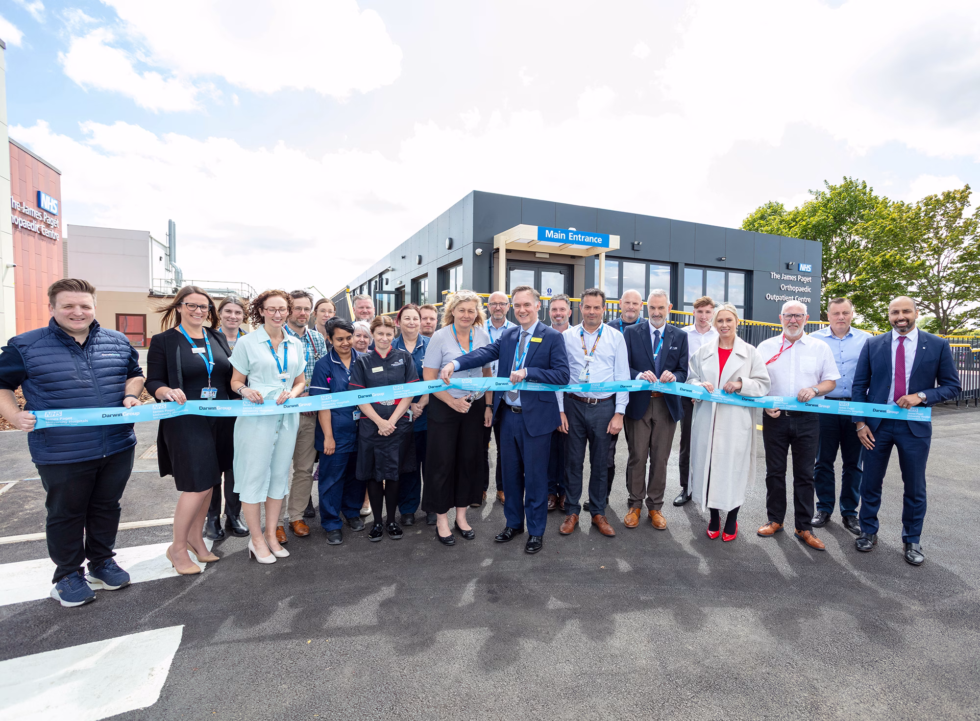 James Paget Orthopaedic Outpatient Centre at Great Yarmouth. Ribbon cutting. A group of clinicians and some members of the project team are stood outside with the building in the background. They are holding a blue ribbon and a lady in the middle of the group is holding a large pair of ceremonial scissors.